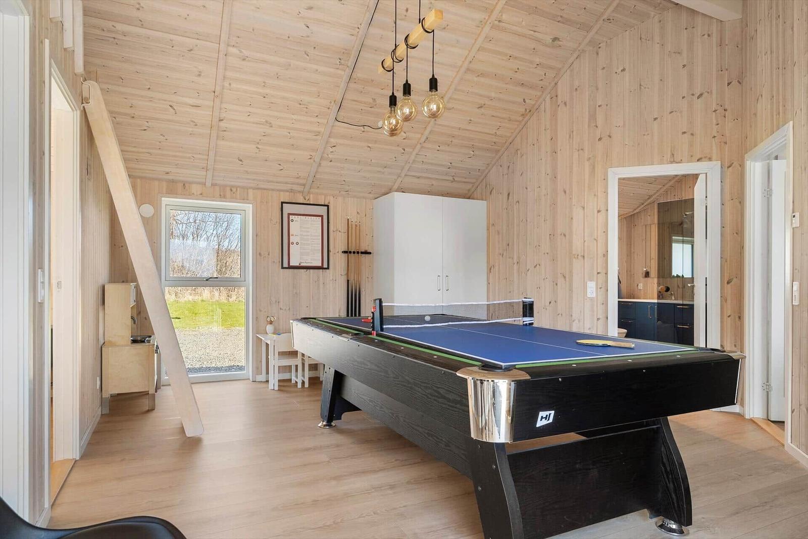 Room with ping pong table, wooden walls, and window view of green area.