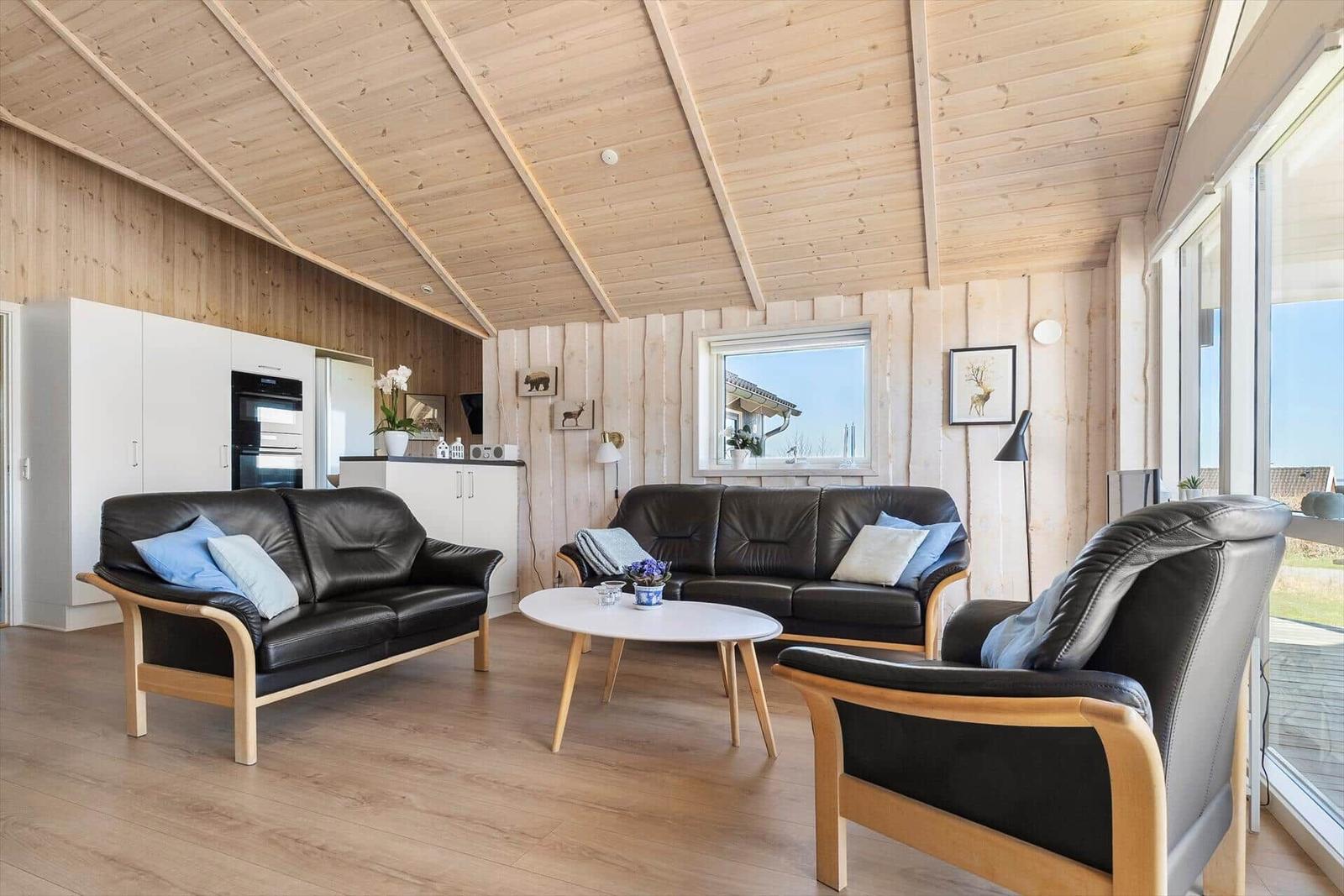 Living room with black leather furniture and wooden wall cladding. Large windows and kitchen area visible.