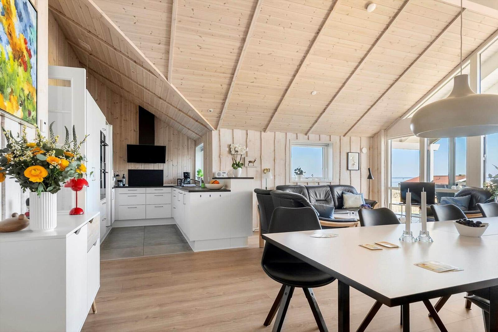 Living and dining area with kitchen under wooden ceiling. Dining table, sofa, and windows with sea view.