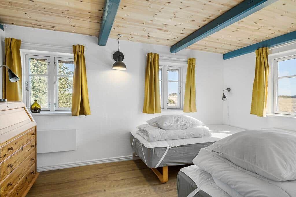 Zwei Betten, Fenster mit gelben Vorhängen, Holzdecke mit blauen Balken.