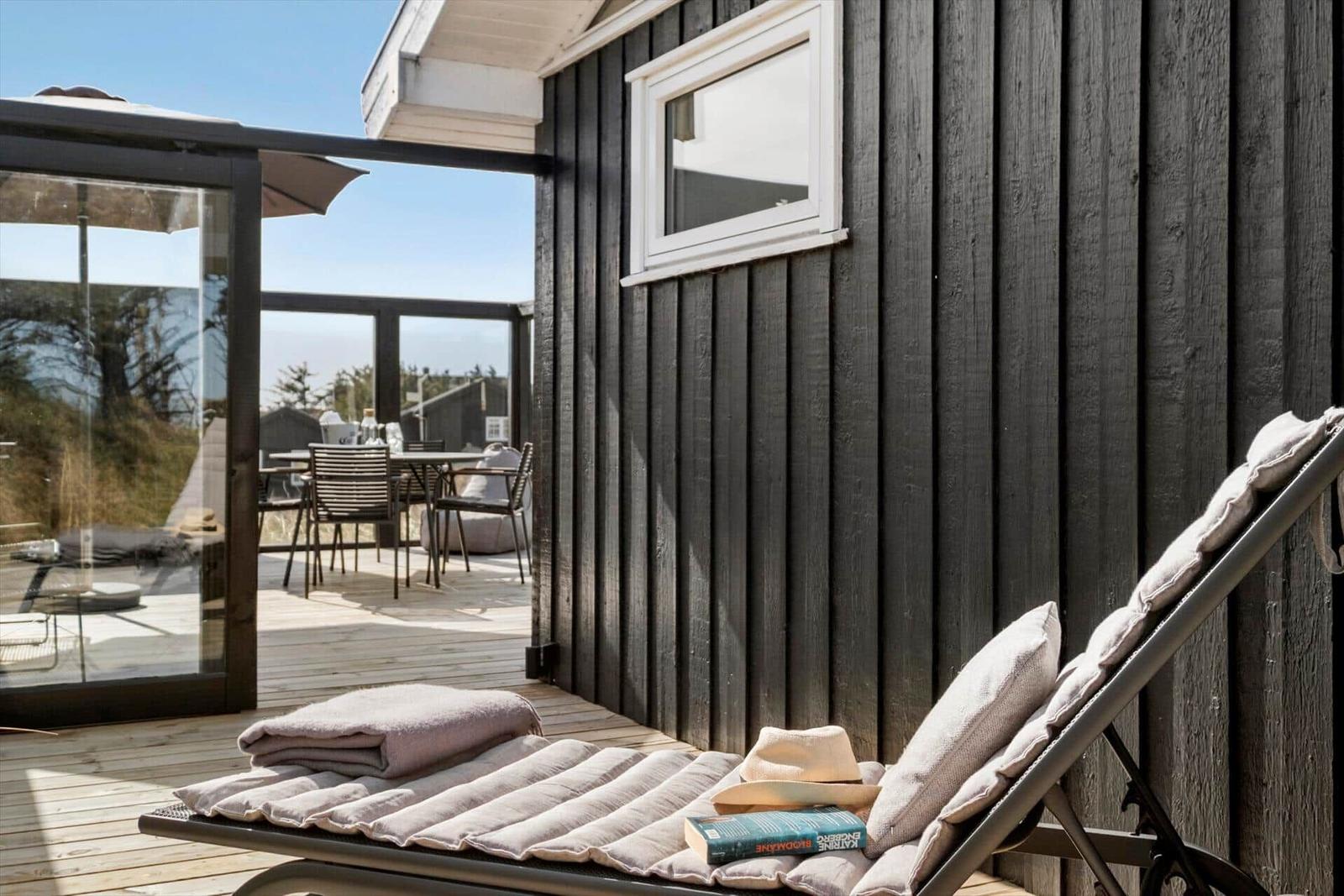 Liegestuhl auf Terrasse neben Haus mit dunkler Fassade und Sicht auf Gartenbereich.