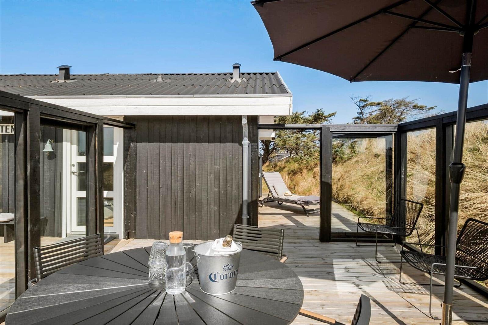 Terrasse mit Tisch, Stühlen und Sonnenschutz. Hintergrund: Haus und Strandvegetation.