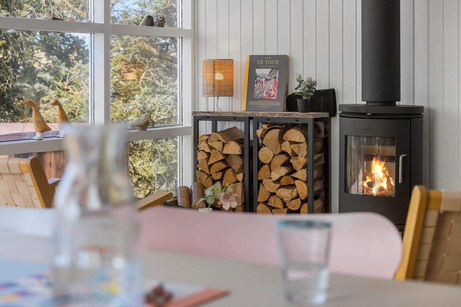 Ein Kamin mit brennendem Holz, daneben ein Holzstapel und ein Fenster mit Blick auf Grün.