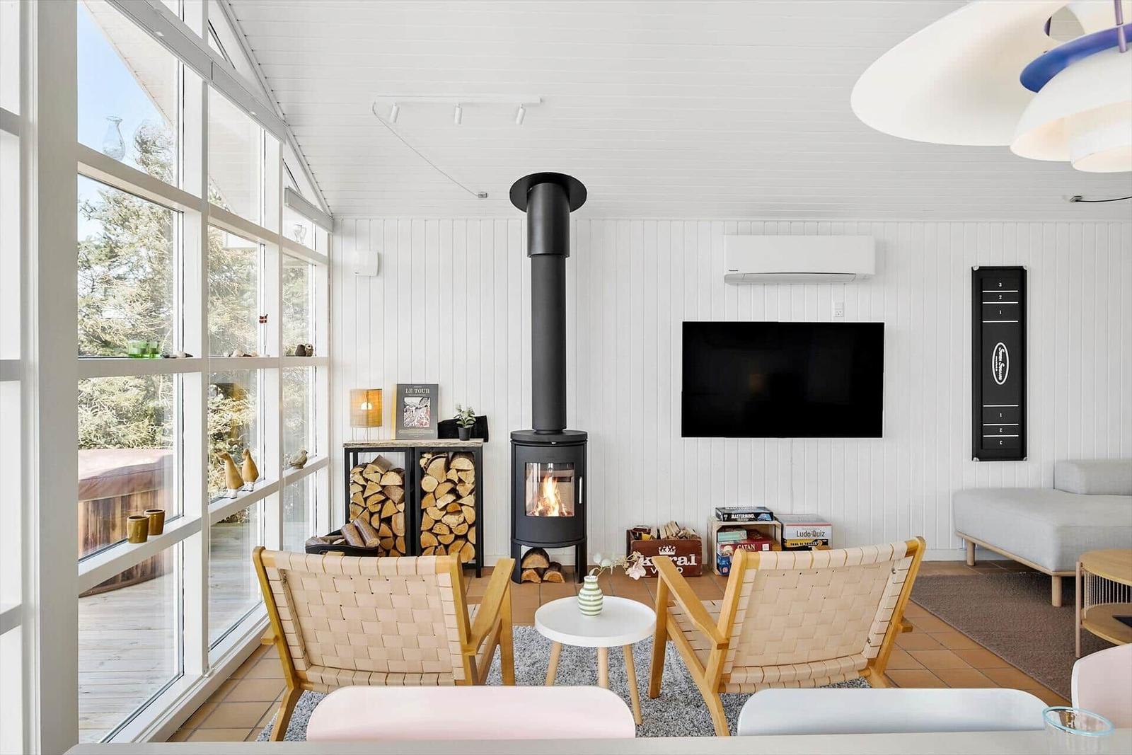 Wohnraum mit Kamin, Fernseher und großen Fenstern. Möbel aus Holz und Stoff.