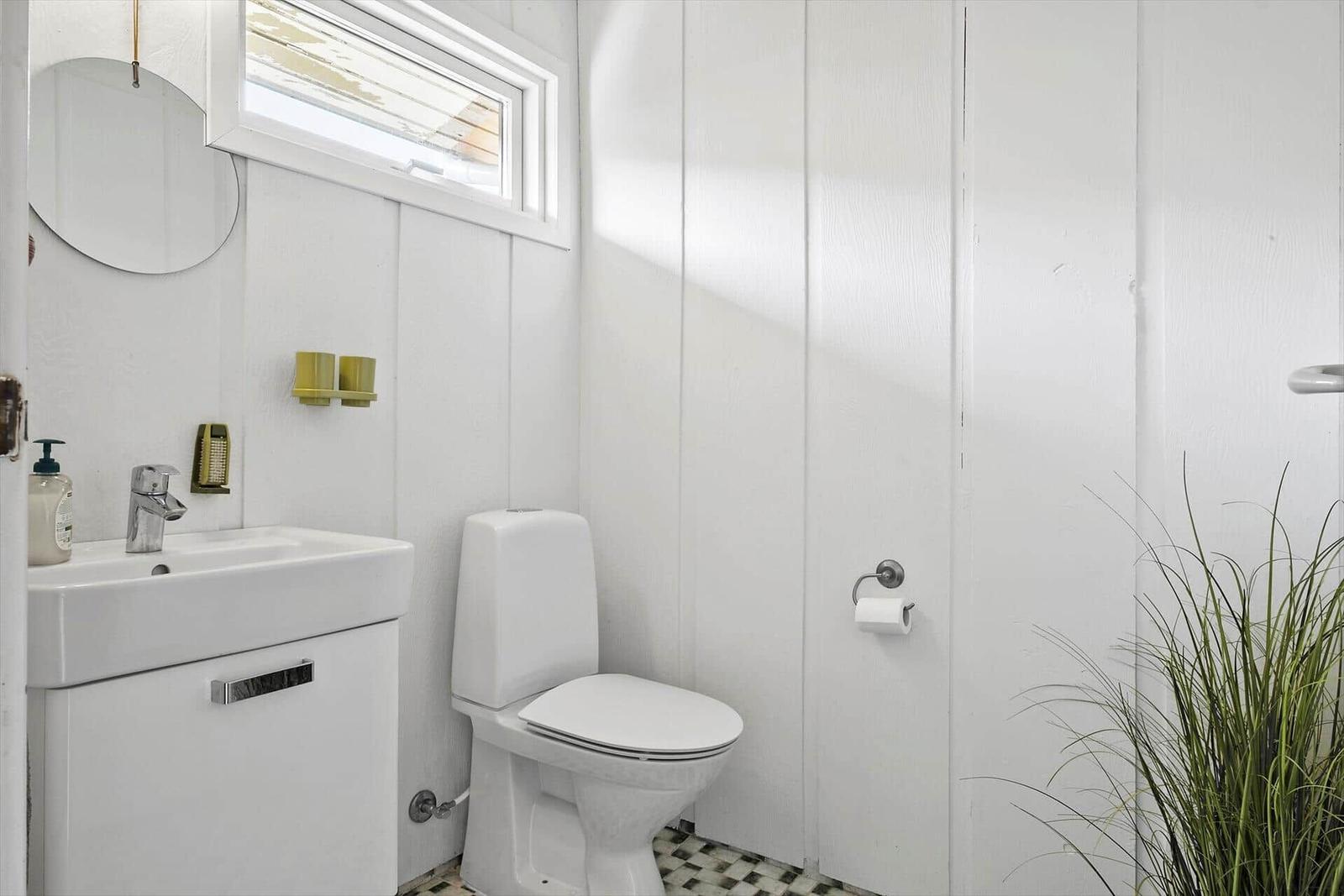 Badezimmer mit Toilette, Waschbecken und Pflanze. Weiße Wände und Bodenfliesen.