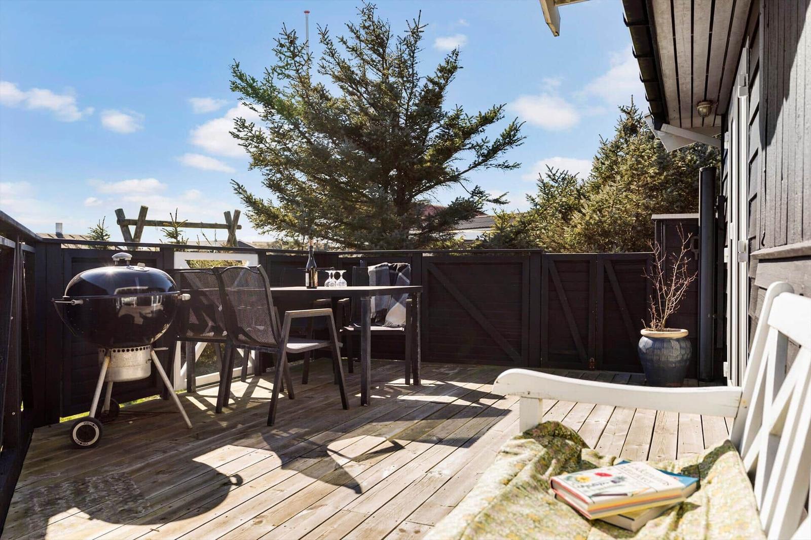 Terrasse mit Grill, Tisch, Stühlen und weißem Liegestuhl. Hintergrund: Holzgeländer und Bäume.