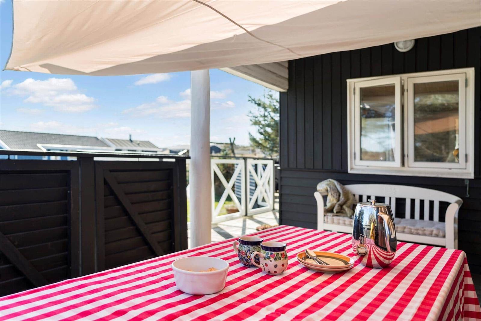 Tisch mit Tischdecke, Tassen und Kanne unter Markise auf Terrasse.