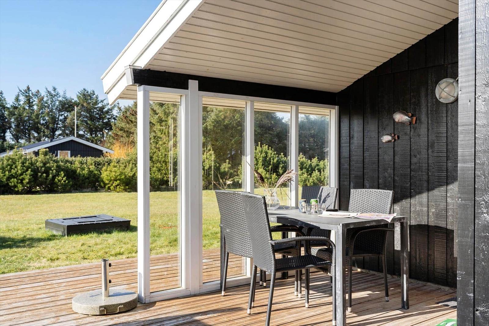 Terrace with table and chairs, glass wall and black wooden wall. Grass and trees in the background.