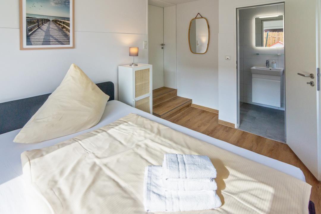 Schlafzimmer mit Bett, Nachttisch und Bad. Holzdielen und Wandspiegel sind sichtbar.