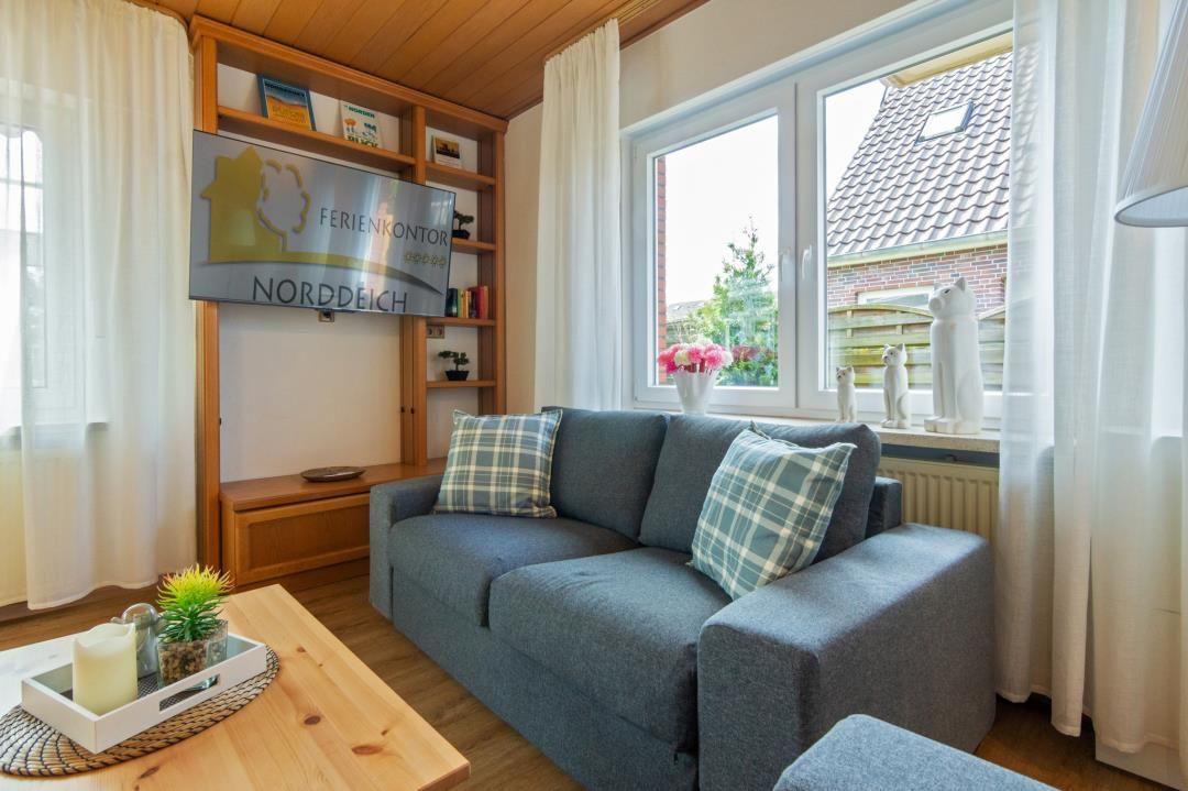 Ein Wohnzimmer mit grauem Sofa, Holztisch und TV. Fenster mit Blick auf Garten.