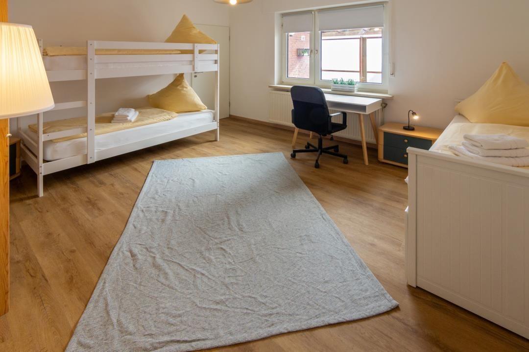 Zimmer mit Doppelstockbett, Einzelbett, Schreibtisch und Fenster.