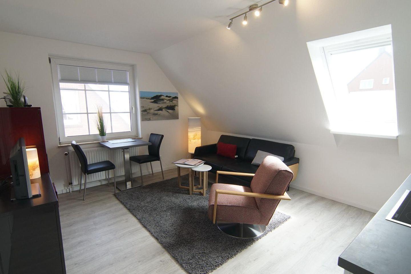 Wohnraum mit Sofa, Sessel, Tisch und Fenstern.