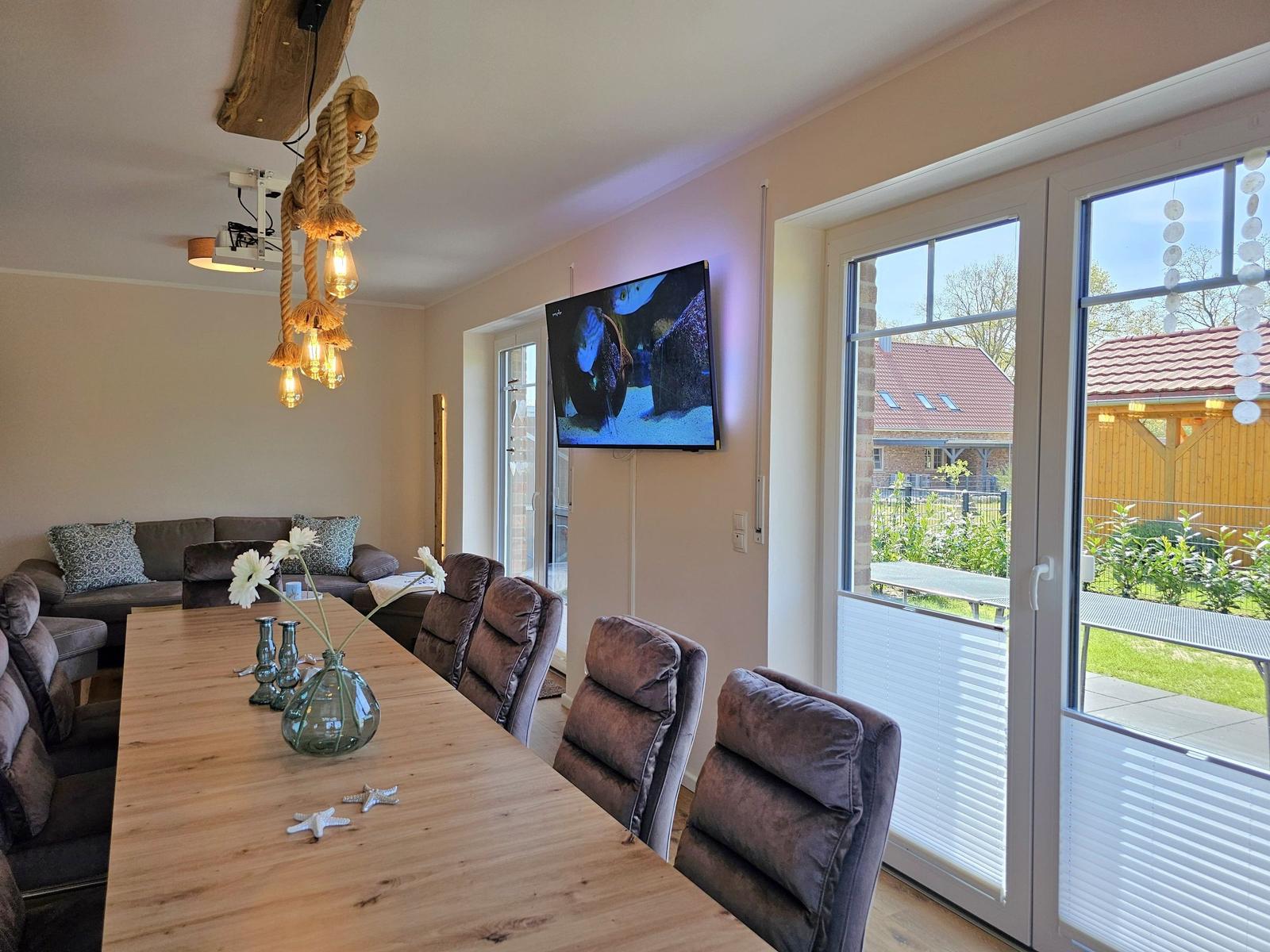 Esszimmer mit Holztisch, Sitzgelegenheiten und TV an der Wand. Blick auf den Garten durch Glastüren.