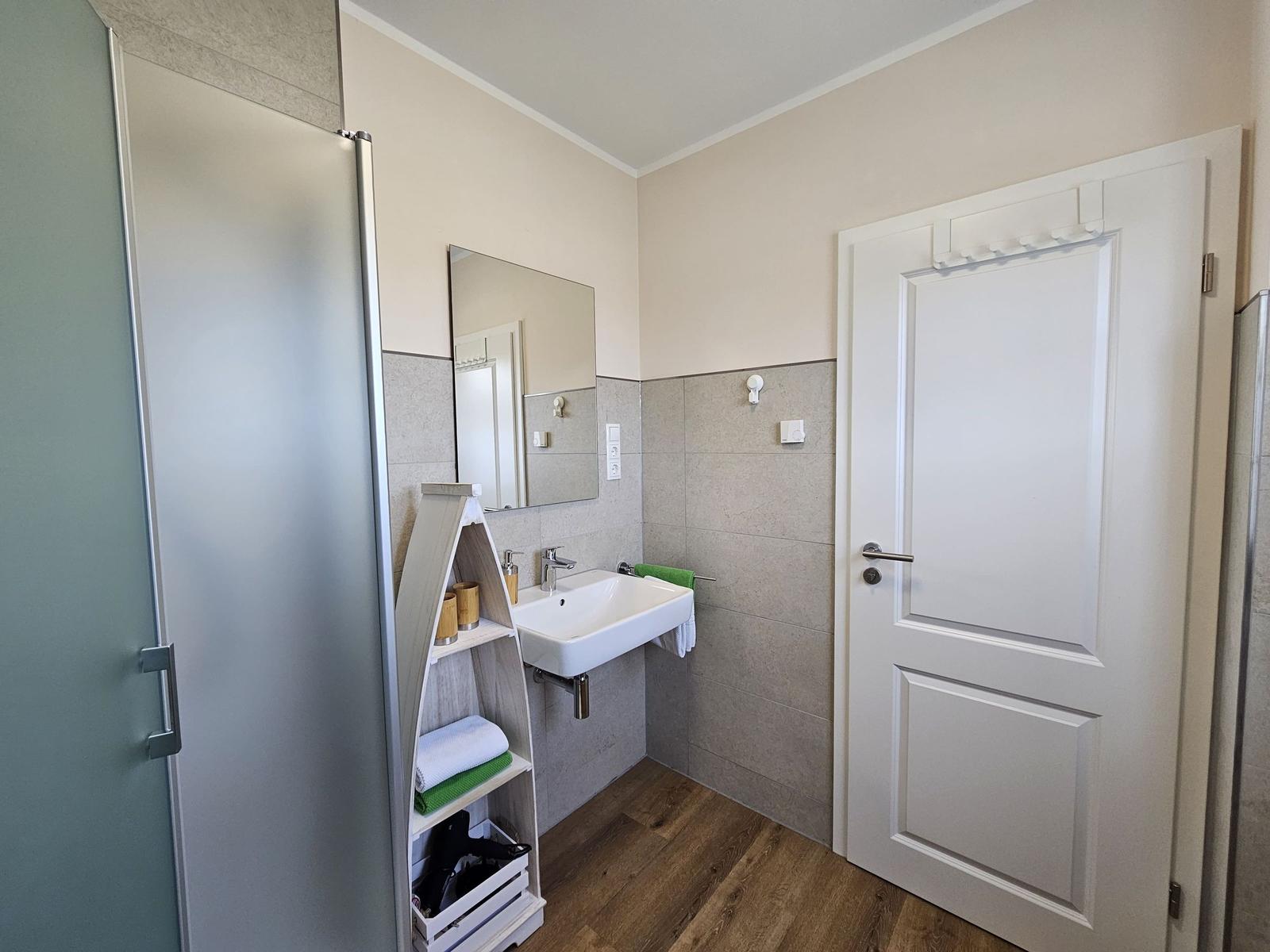 Badezimmer mit Dusche, Waschbecken und Spiegel. Holzdielenboden und graue Fliesen.