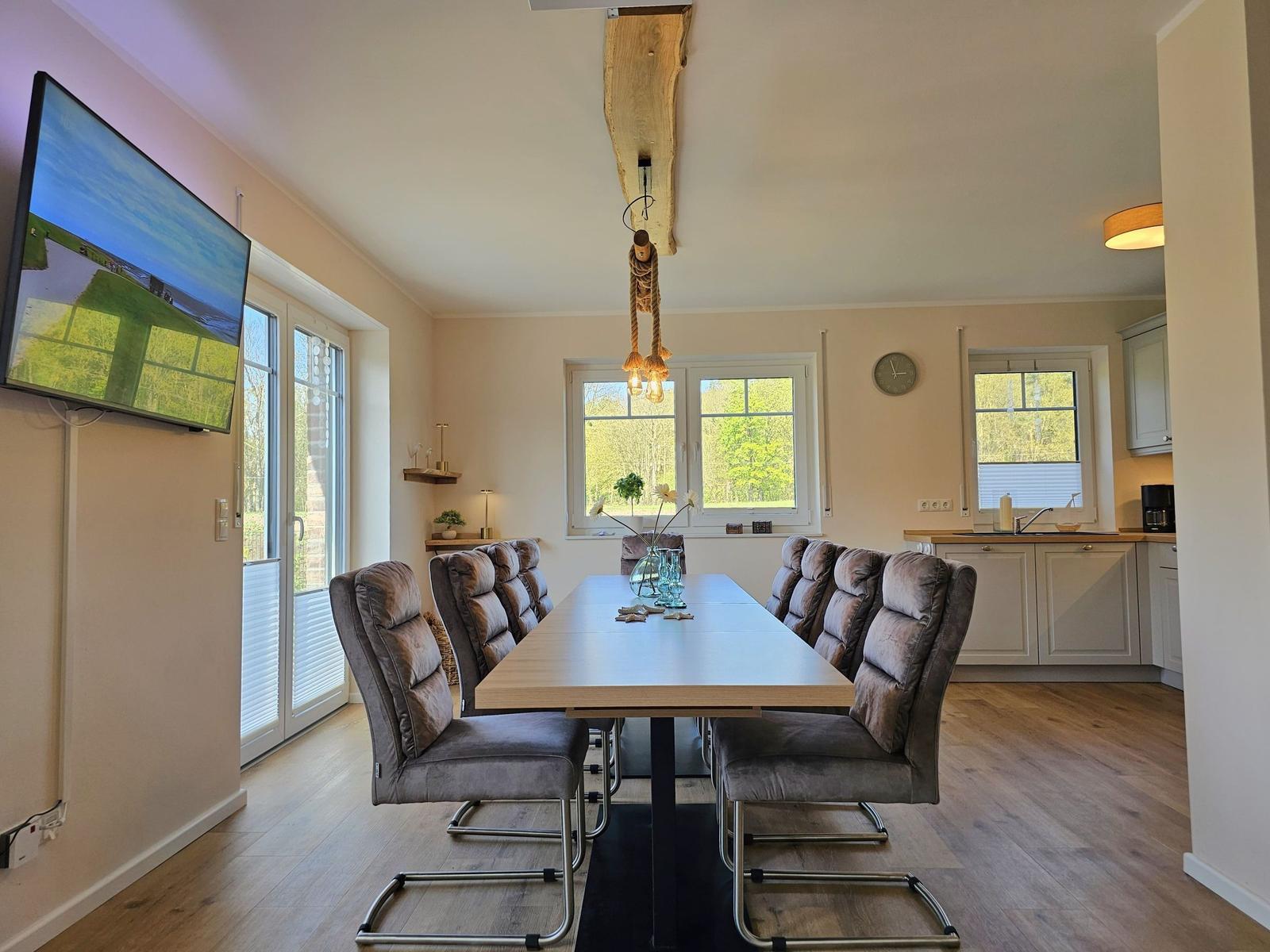 Esszimmer mit langem Tisch, sechs Stühlen und Küchenbereich im Hintergrund.