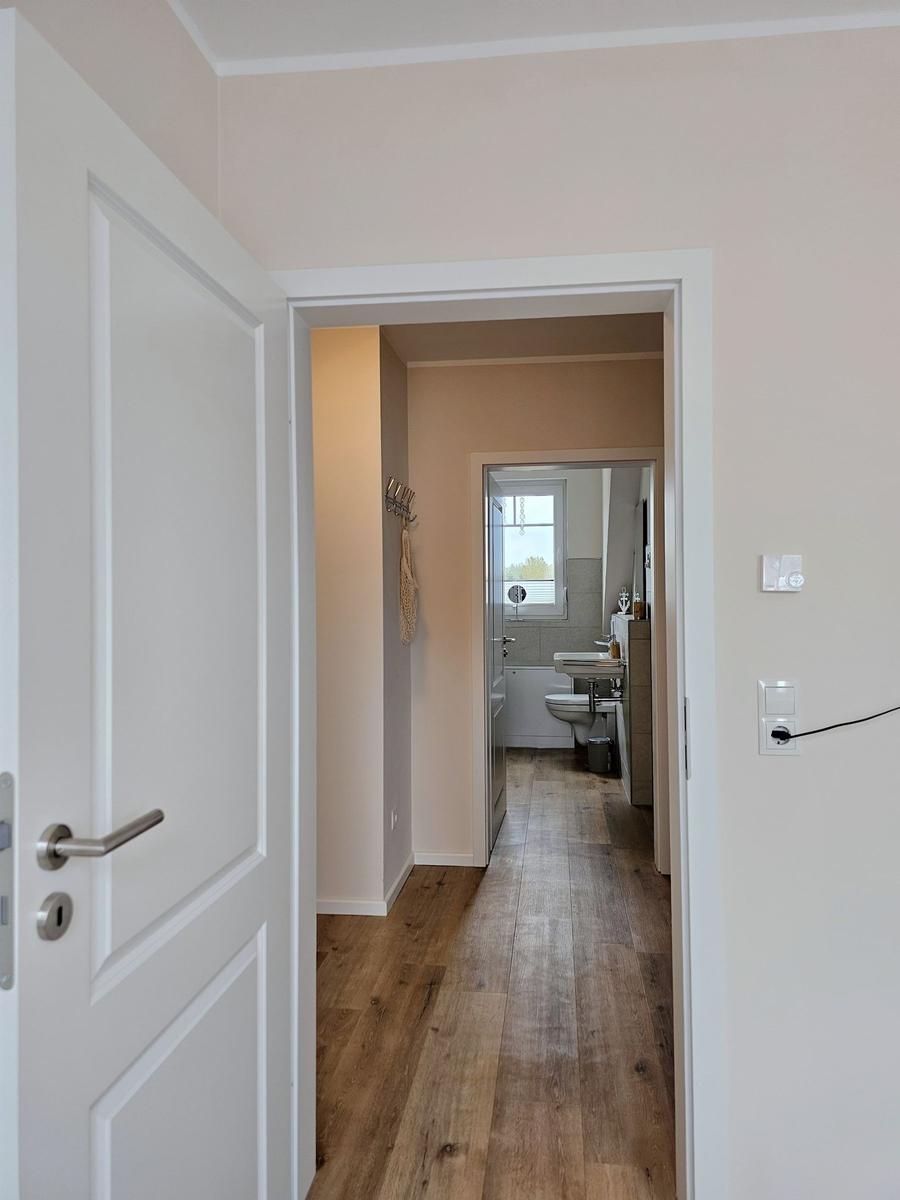 Flur mit Holzboden führt in das Badezimmer. Weiße Türen und Wände.