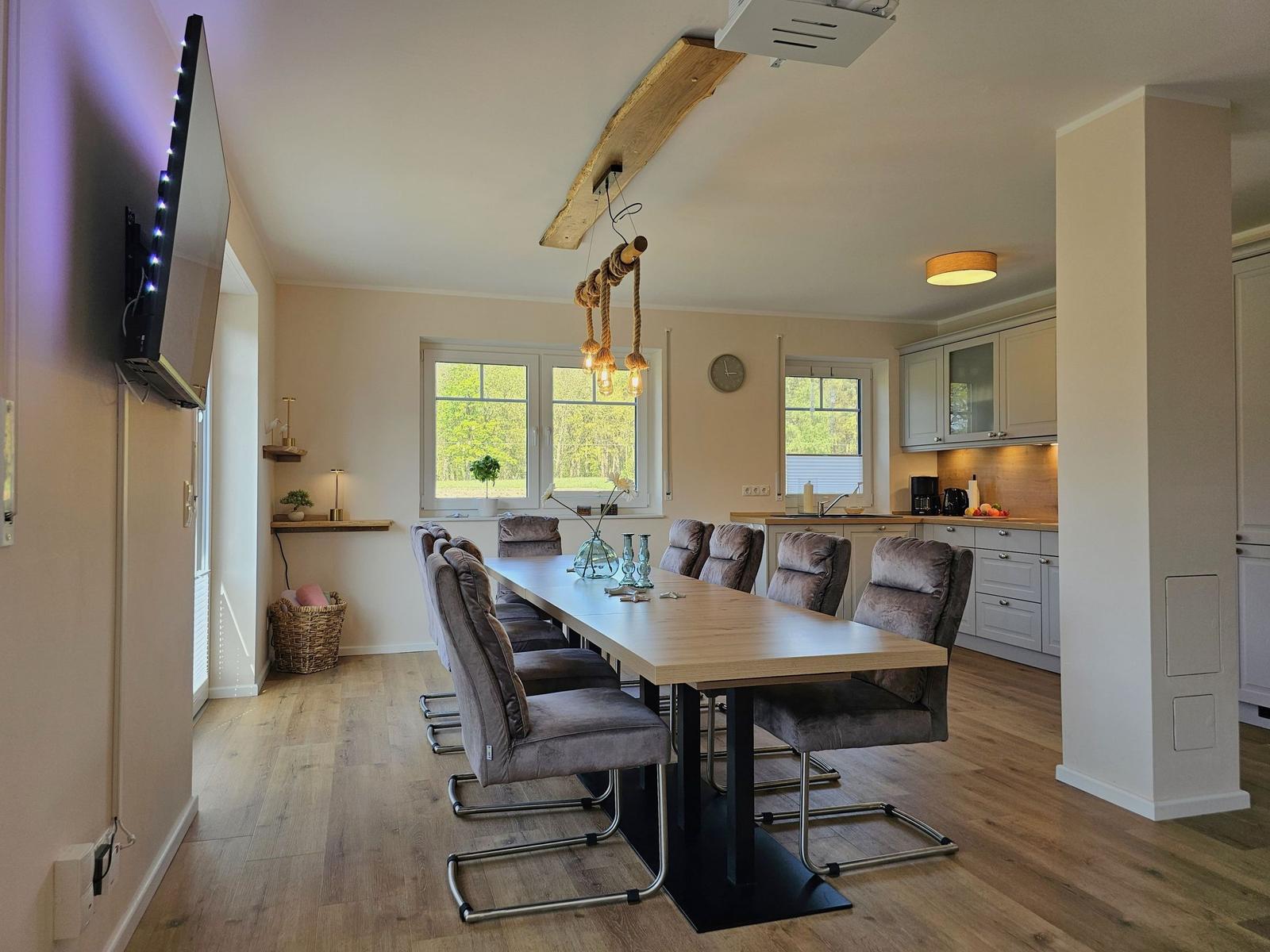 Essbereich mit langer Tisch und Stühlen, Küchenbereich im Hintergrund.