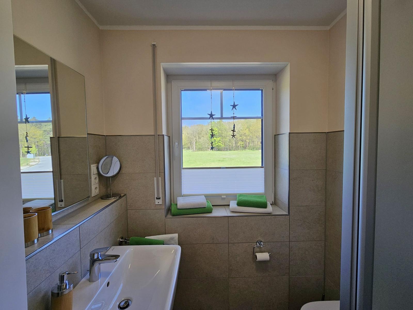 Badezimmer mit Waschbecken, Spiegel und Fenster mit Blick auf Grünfläche.