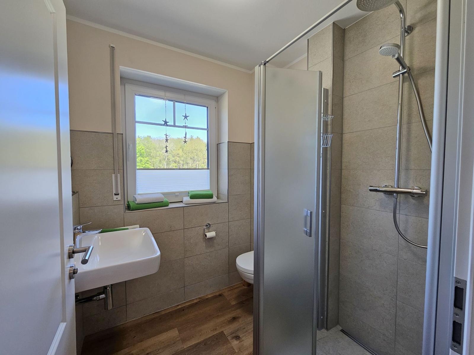 Bad mit Dusche, Waschbecken und Toilette. Fenster mit Blick auf Wald.