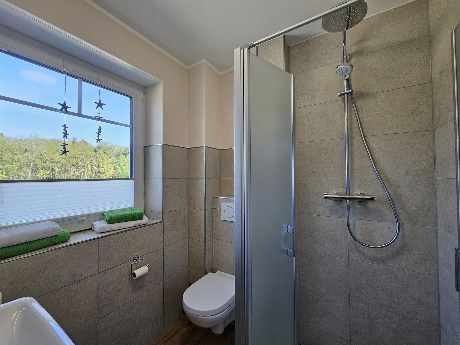 Badezimmer mit Dusche, Toilette und Fenster mit Blick auf Wald.
