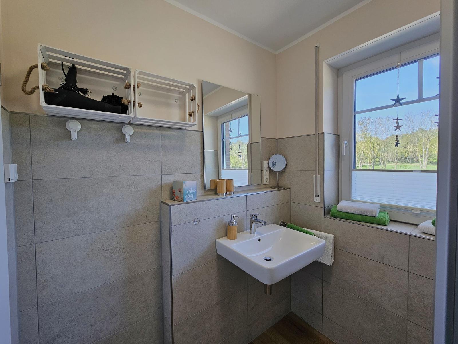 Badezimmer mit Waschbecken, Spiegel und Fenster mit Blick auf Grünfläche.