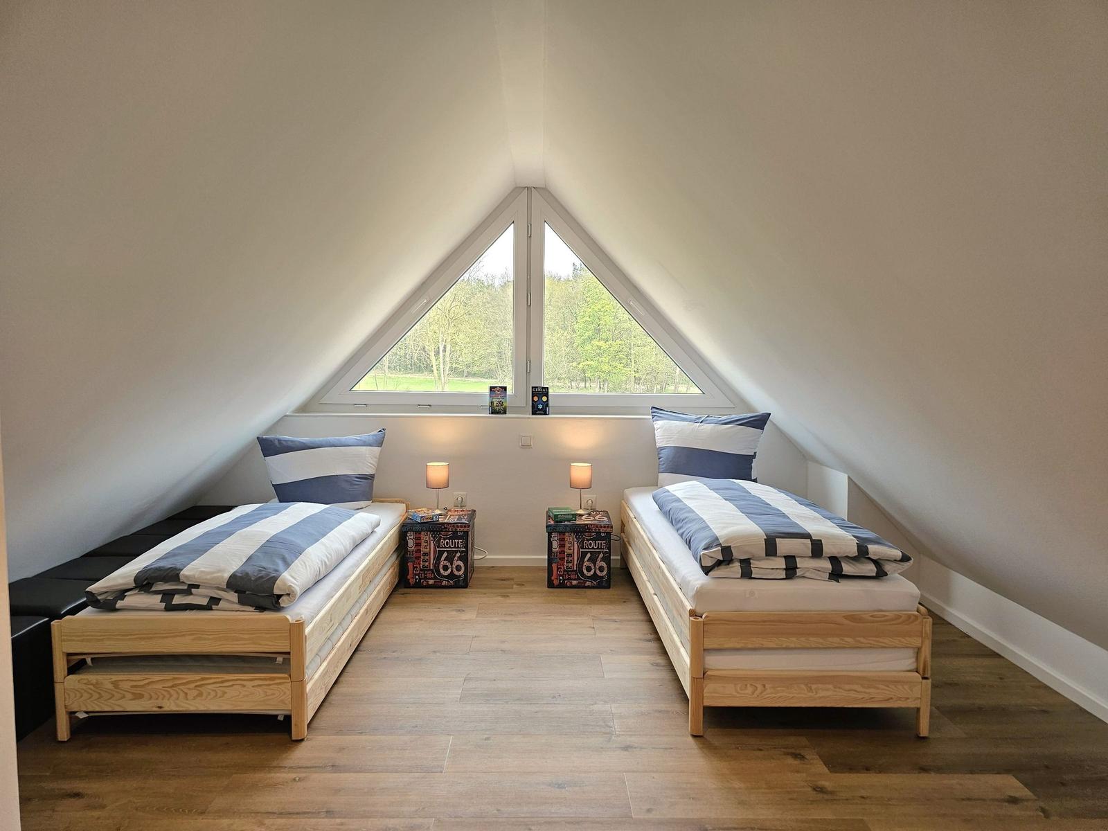 Zwei Betten unter einem Dachfenster mit Blick auf einen Wald.