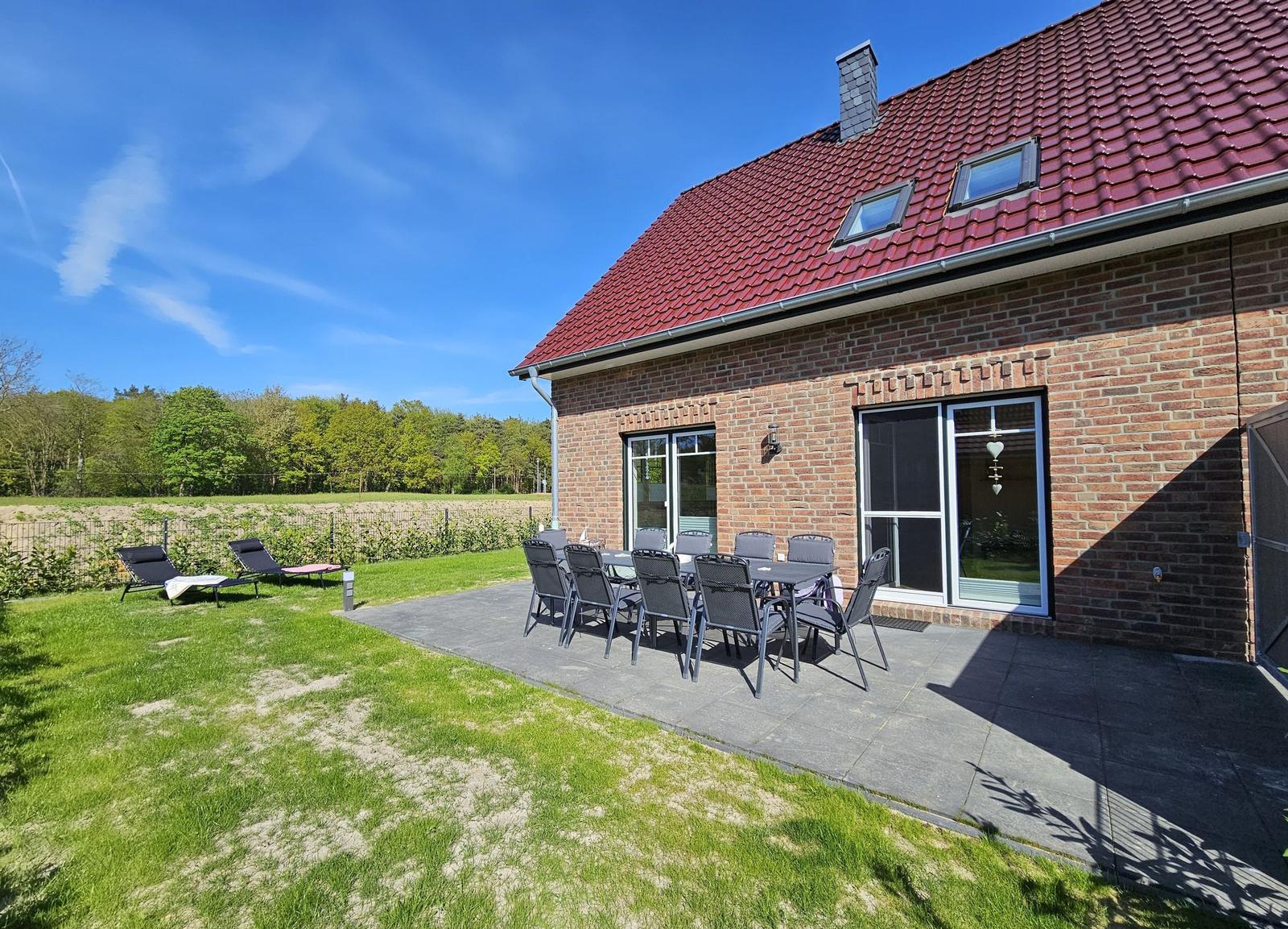 Haus mit Terrasse, Garten und Außentisch mit Stühlen. Hintergrund: Wald und blauer Himmel.