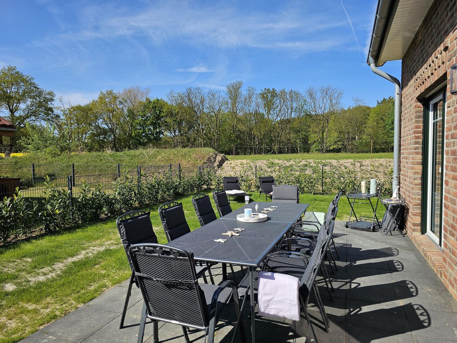 Terrasse mit Tisch und Stühlen, Grünfläche und Wald im Hintergrund.