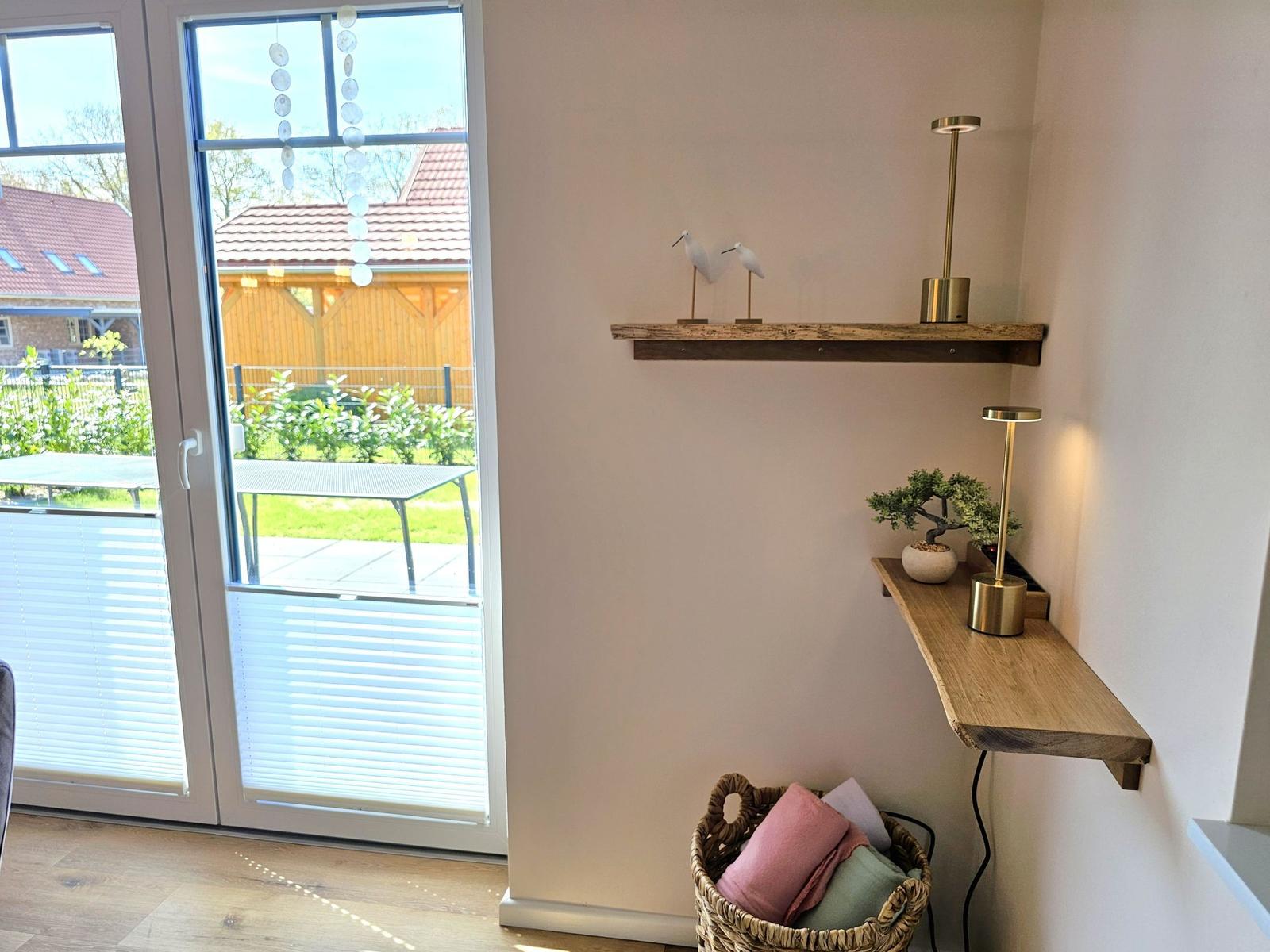 Eingangsbereich mit Holzregalen, Lampe und Korb mit Kissen. Durchgang zur Terrasse mit Garten.