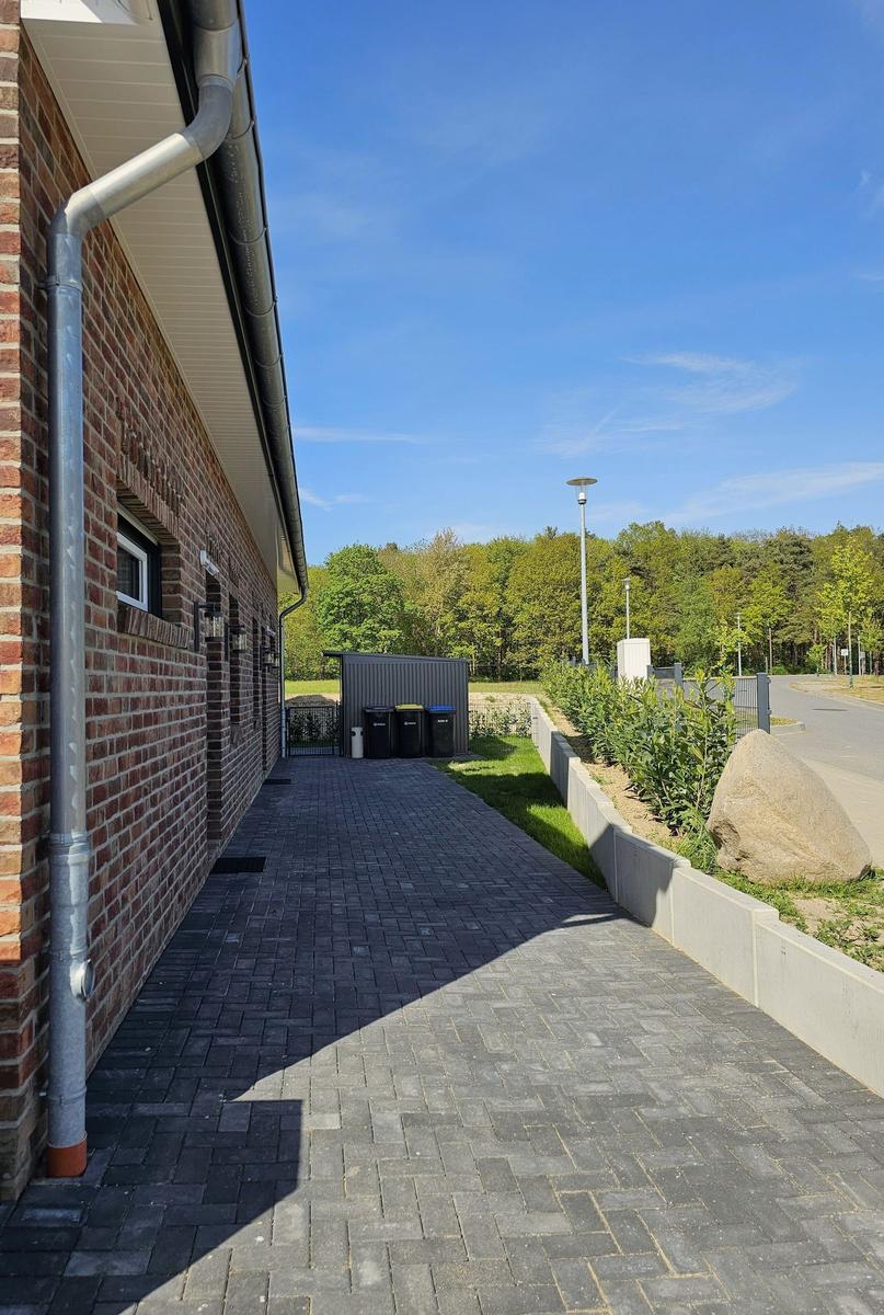 Pflasterweg neben Backsteinhaus mit Garten und Wald im Hintergrund.