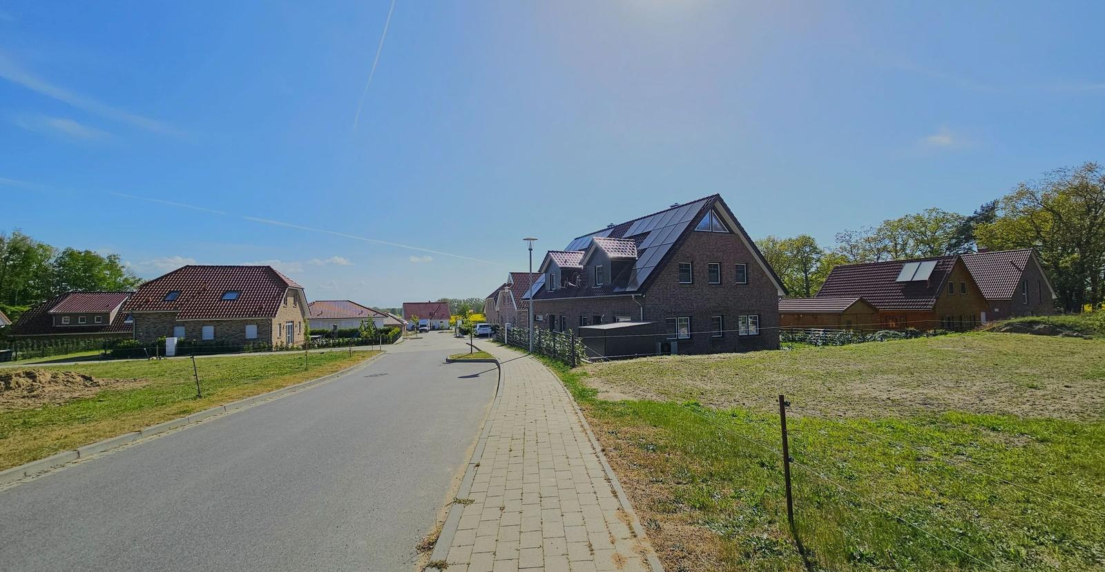 Straße mit Häusern und Grünflächen unter blauem Himmel.