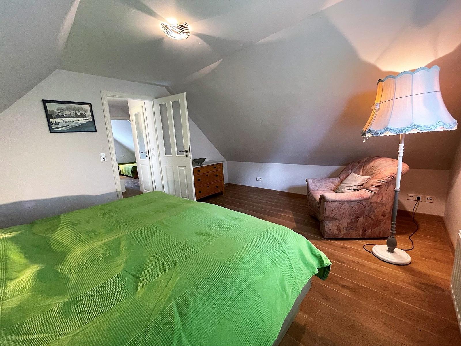 Schlafzimmer mit grünem Bett, Sessel und Stehlampe unter schrägen Dachschrägen.
