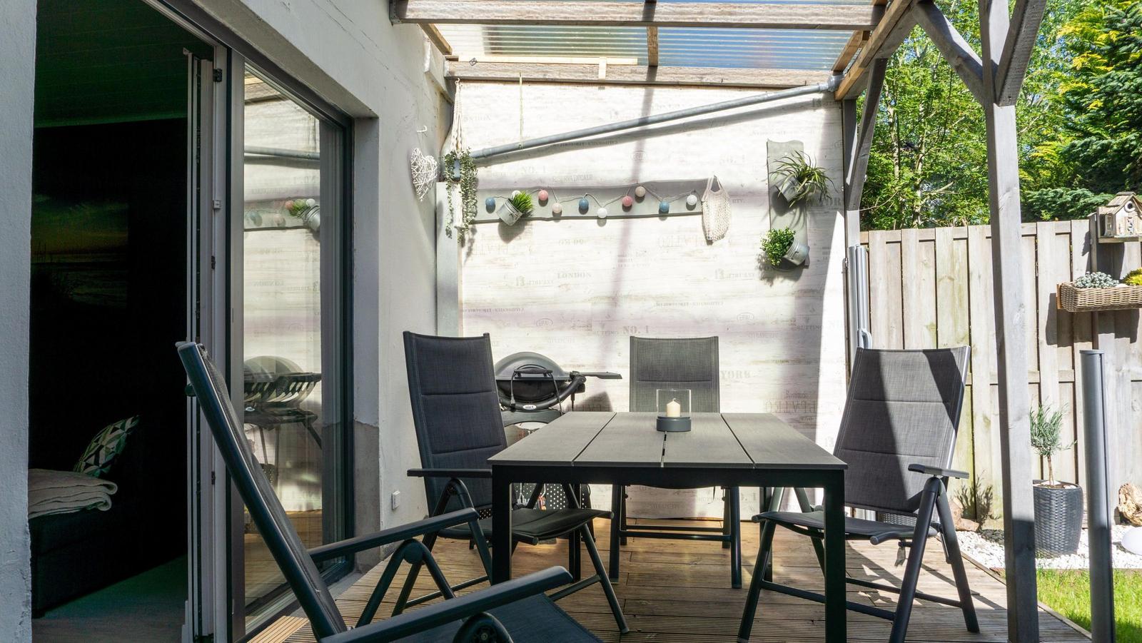 Terrasse mit Tisch, Stühlen und Grill unter Dach. Hintergrund: Holzzaun und Pflanzen.