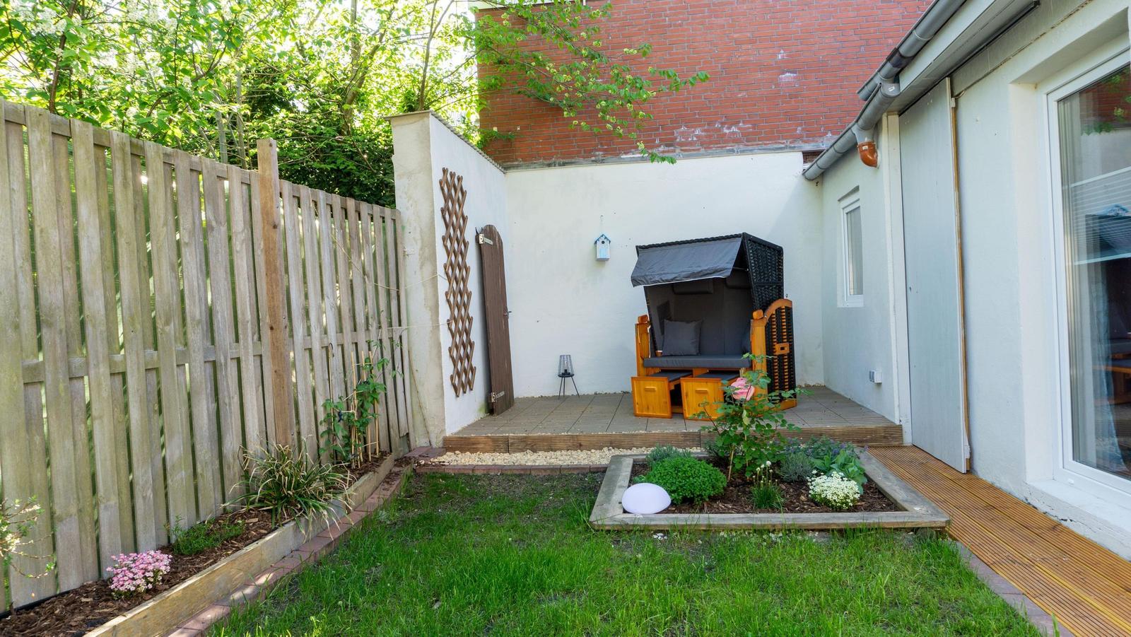 Hinterhof mit Garten, Holzgeländer, Sitzgelegenheit und Blumenbeete.