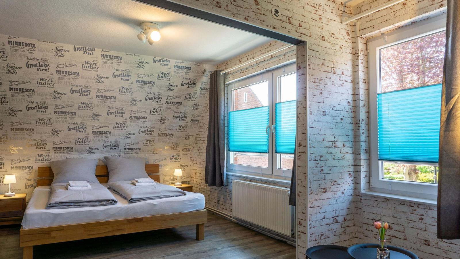 Schlafzimmer mit Doppelbett, Fenstern mit blauen Jalousien und weiß verputzten Ziegelwänden.