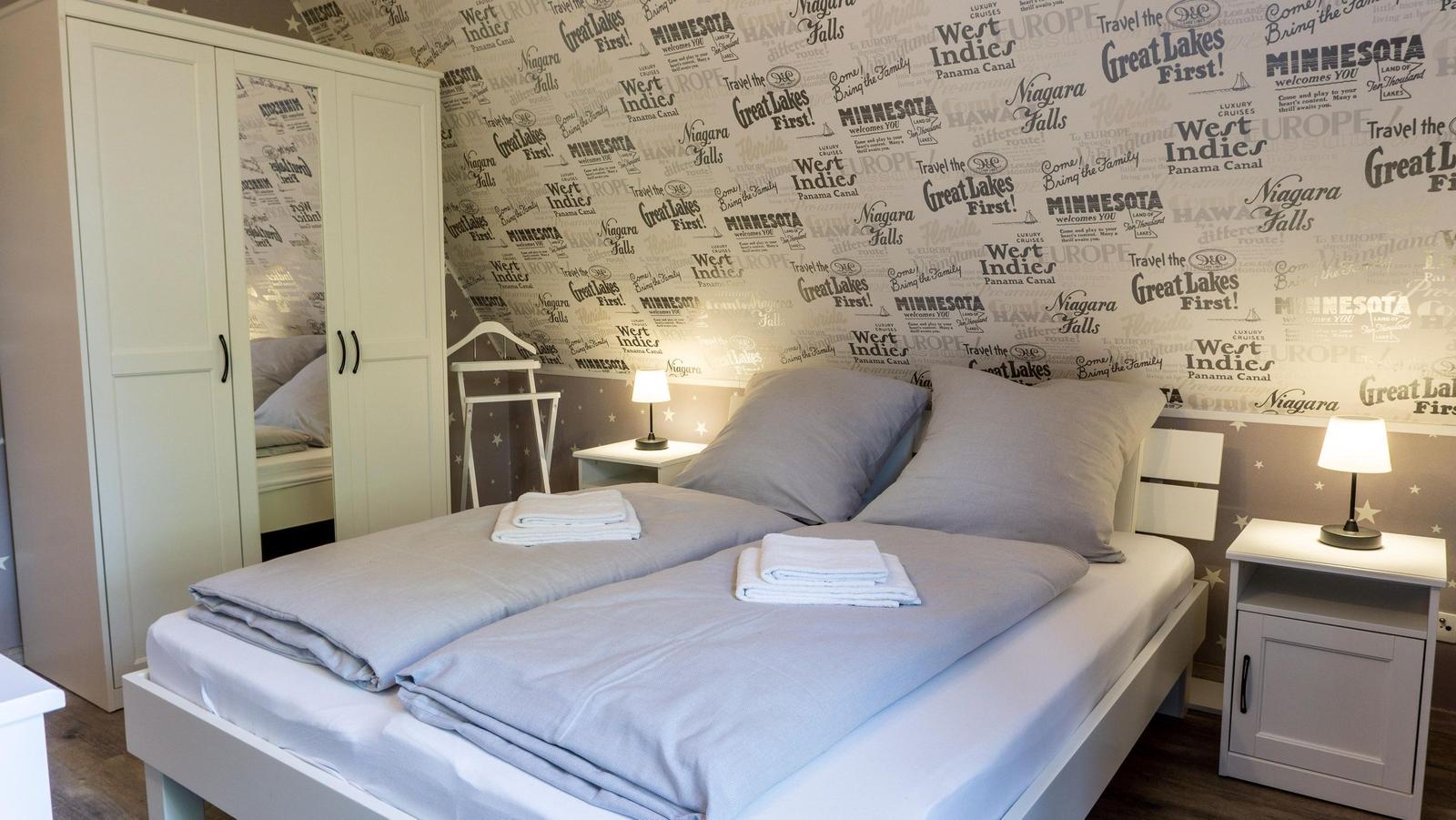 Schlafzimmer mit Doppelbett, zwei Nachttischen und Wandtapete mit Reisestichworten.