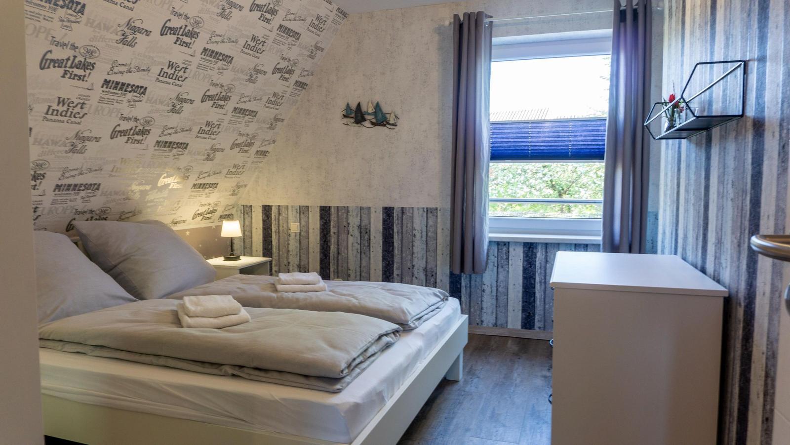 Schlafzimmer mit Doppelbett, Fenster und Schrank. Wand mit Reisemotiv und Holzoptik.