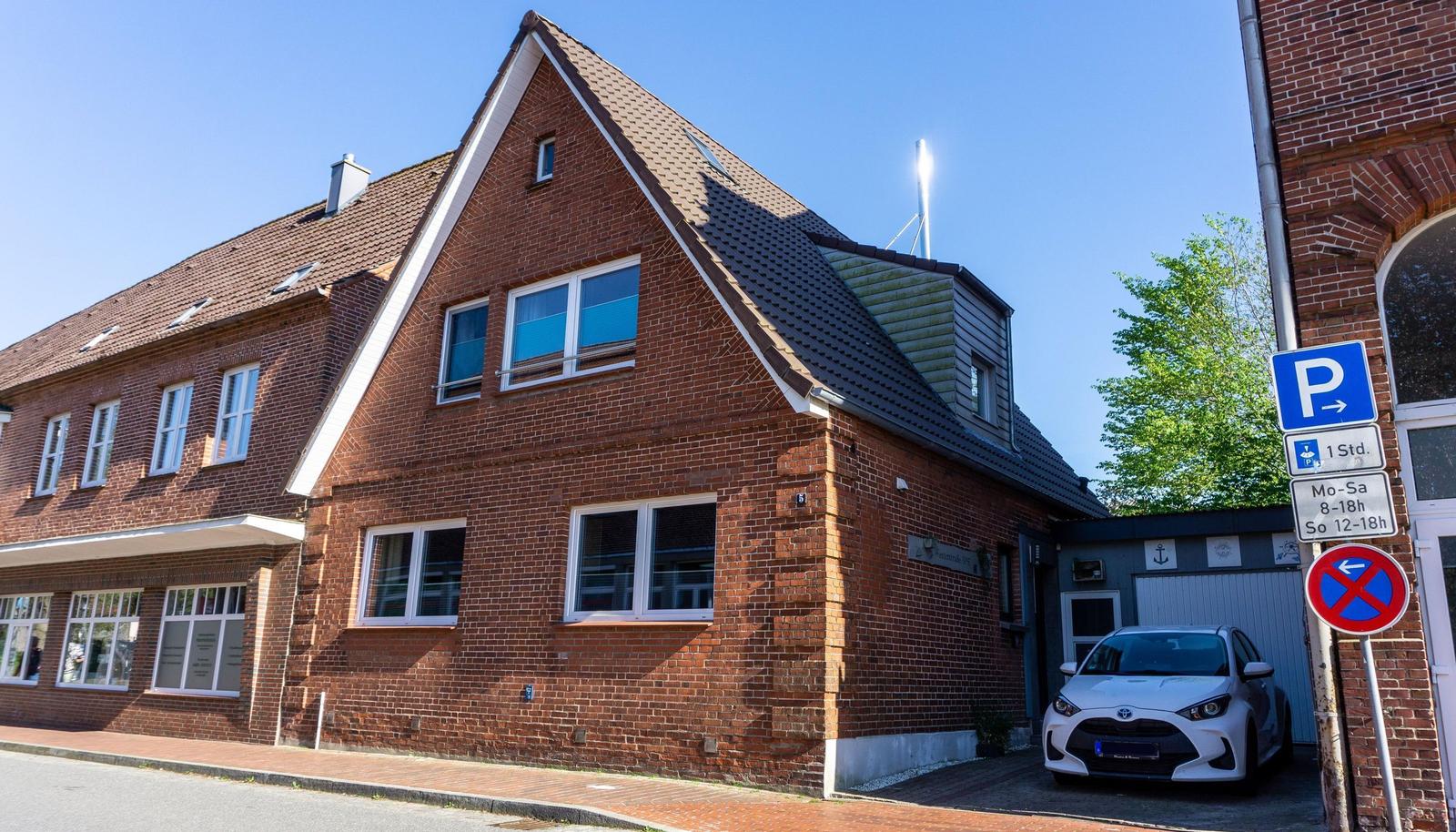 Rotes Backsteingebäude mit Dach und Fenstern, daneben Parkplatz mit Schildern und Auto.
