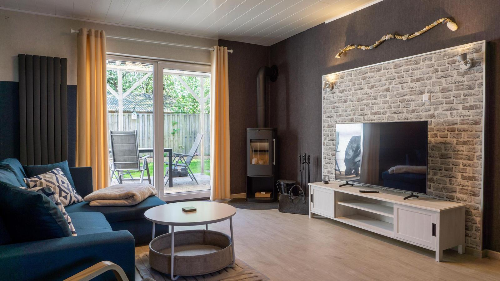 Wohnzimmer mit Sofa, Fernseher und Holzofen. Blick auf Garten durch Glastür.