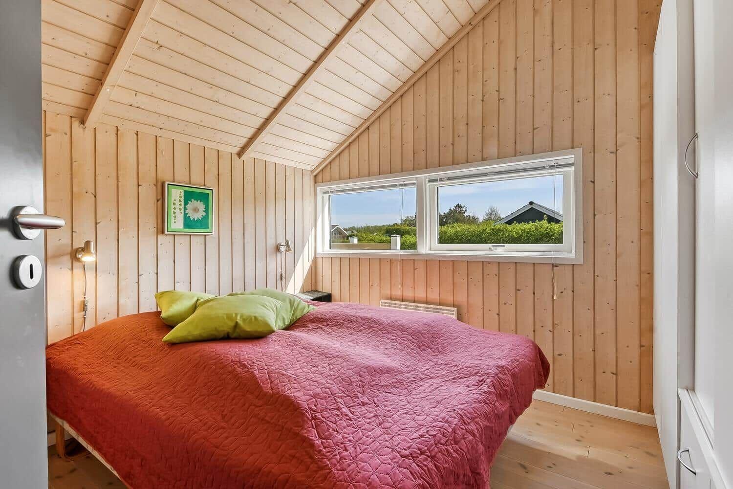 Schlafzimmer mit Holzwand, Bett mit rotem Decken und grünen Kissen, Fenster mit Blick auf Außenbereich.