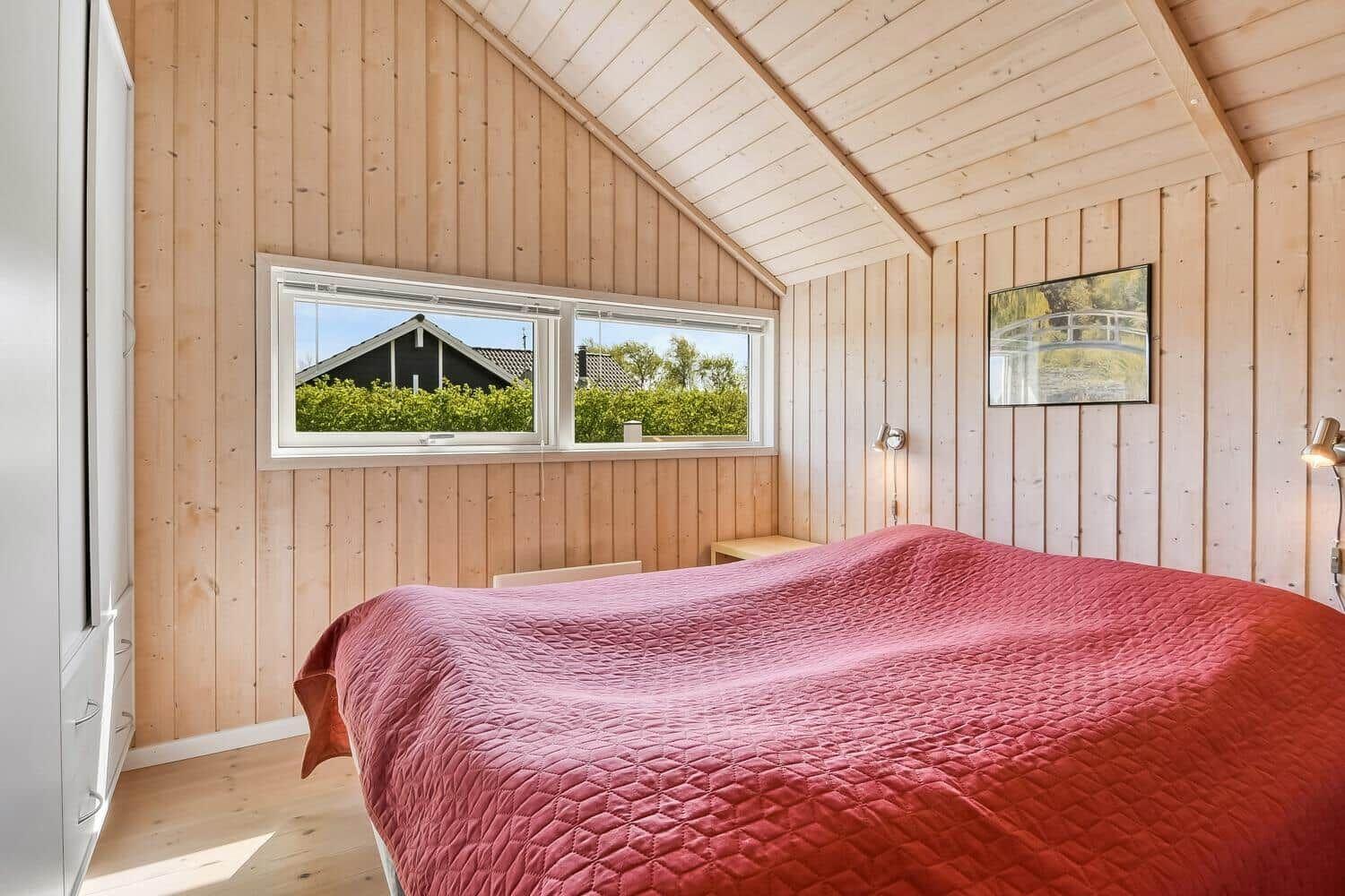 Schlafzimmer mit Holzwand und Dachfenster. Bett mit roter Bettdecke. Wandbild und Nachttische.
