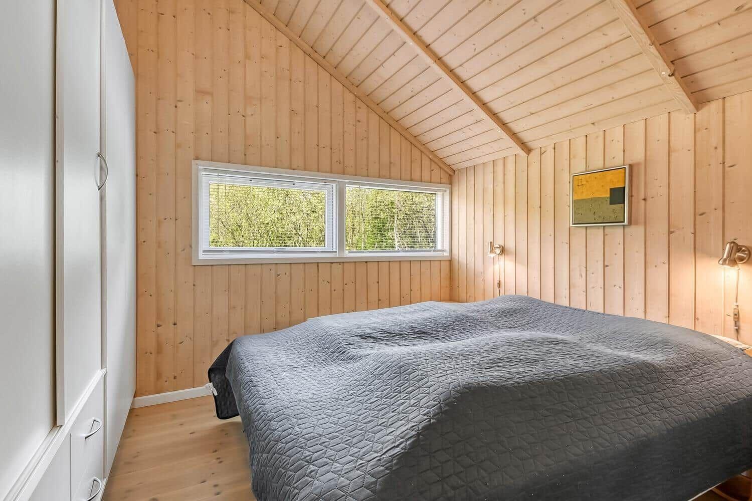 Schlafzimmer mit Holzwand, Doppelbett und Fenster mit Blick auf Bäume.