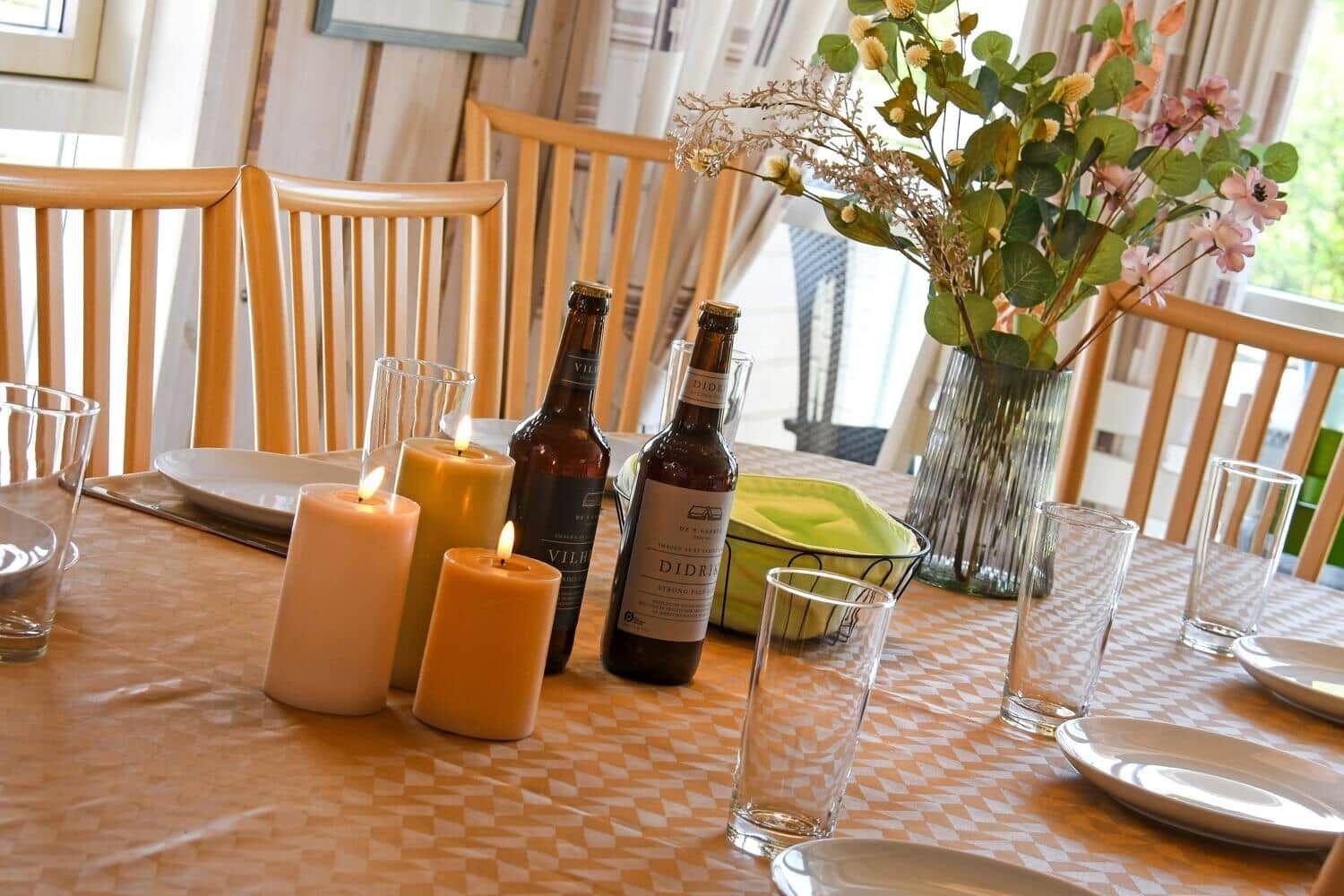 Tisch mit Kerzen, Flaschen und Blumenarrangement. Holzstühle und Tischdecke mit Muster.