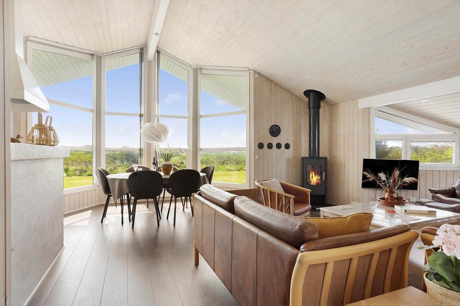 Wohn- und Essbereich mit Holzwand, Kamin und großen Fenstern. Esstisch mit Stühlen und Sofa.