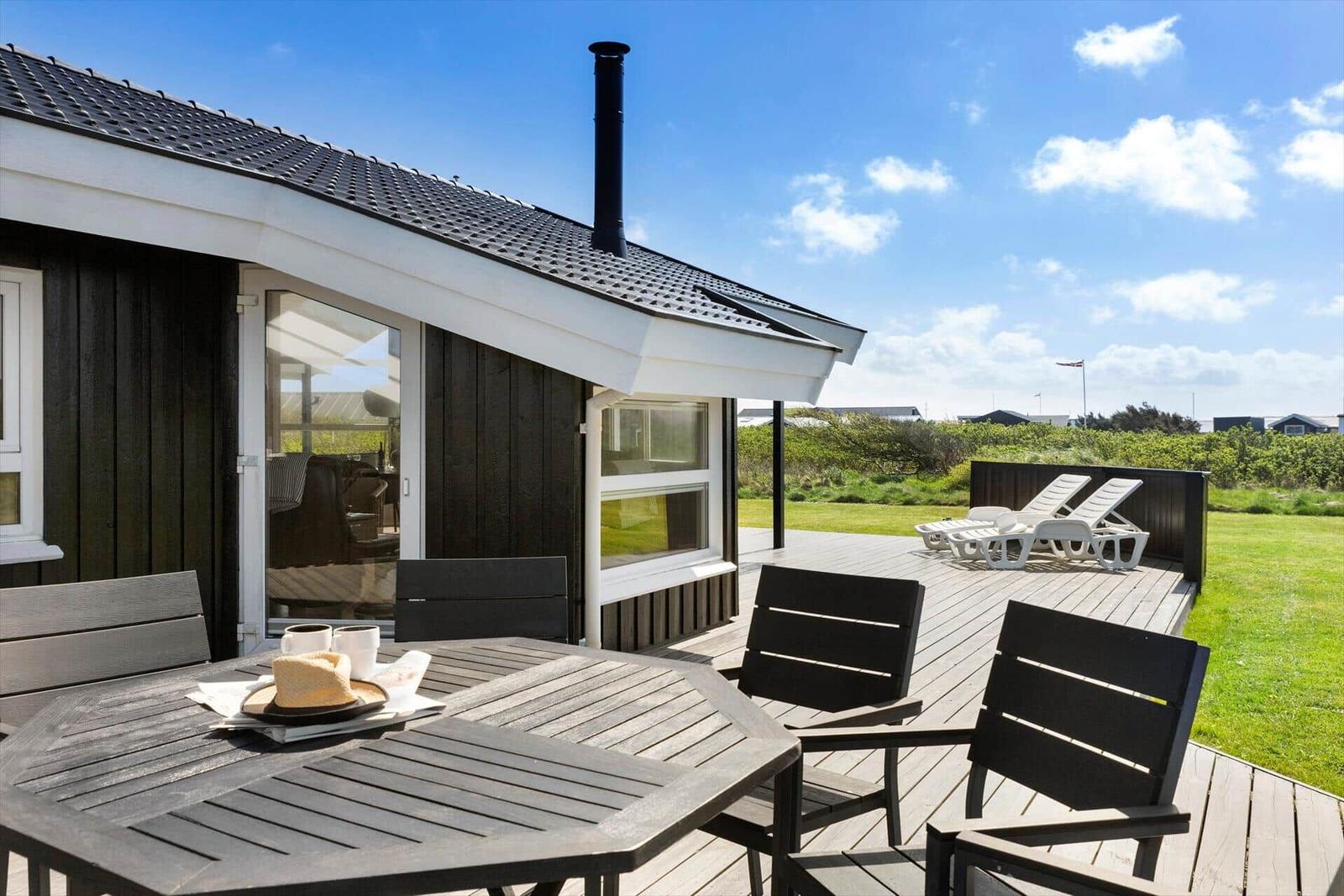 Terrasse mit Tisch, Stühlen und Liegen. Haus mit schwarzem Holz und Dachrohr.