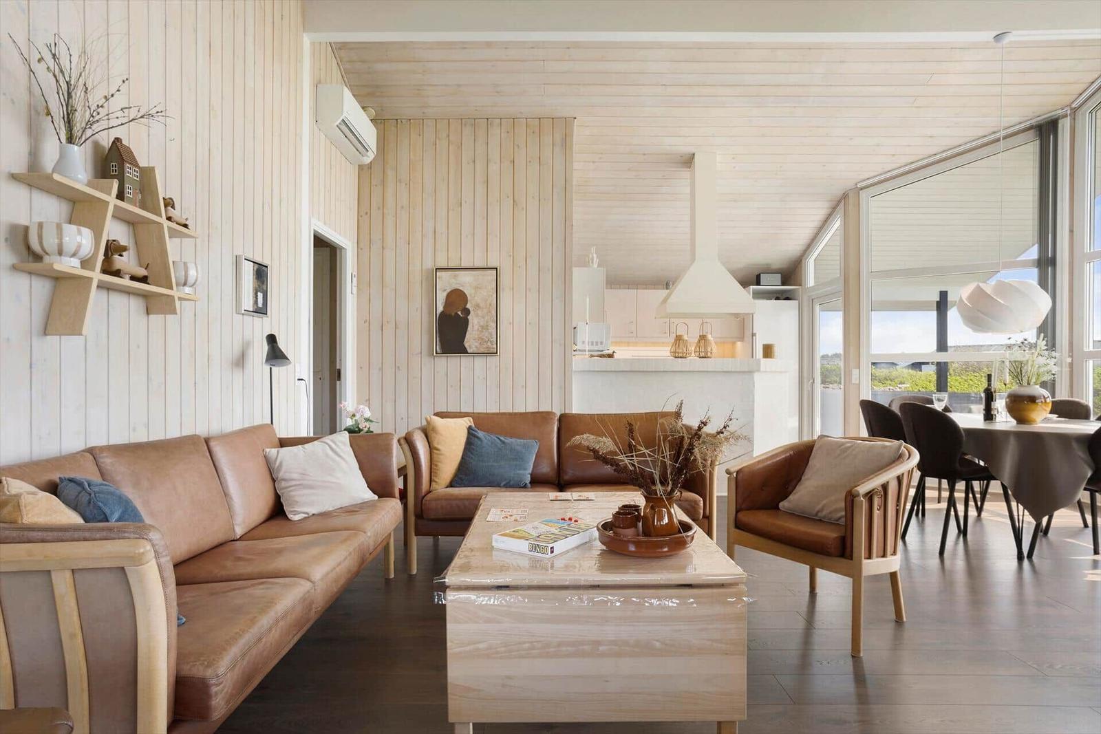 Wohn- und Essbereich mit Holzwand, Sofa und Tisch. Küche im Hintergrund. Große Fenster mit Blick.