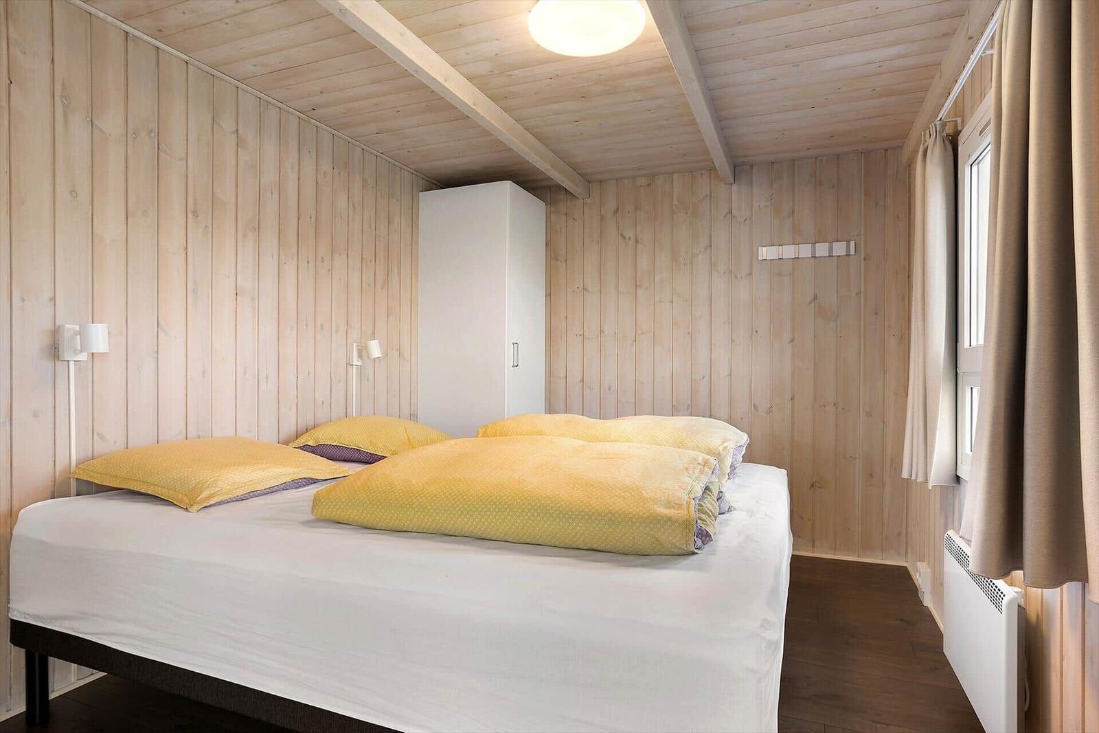 Schlafzimmer mit Holzwand, Bett mit gelben Kissen und weißem Bettzeug, weißer Schrank und Fenster mit Vorhängen.