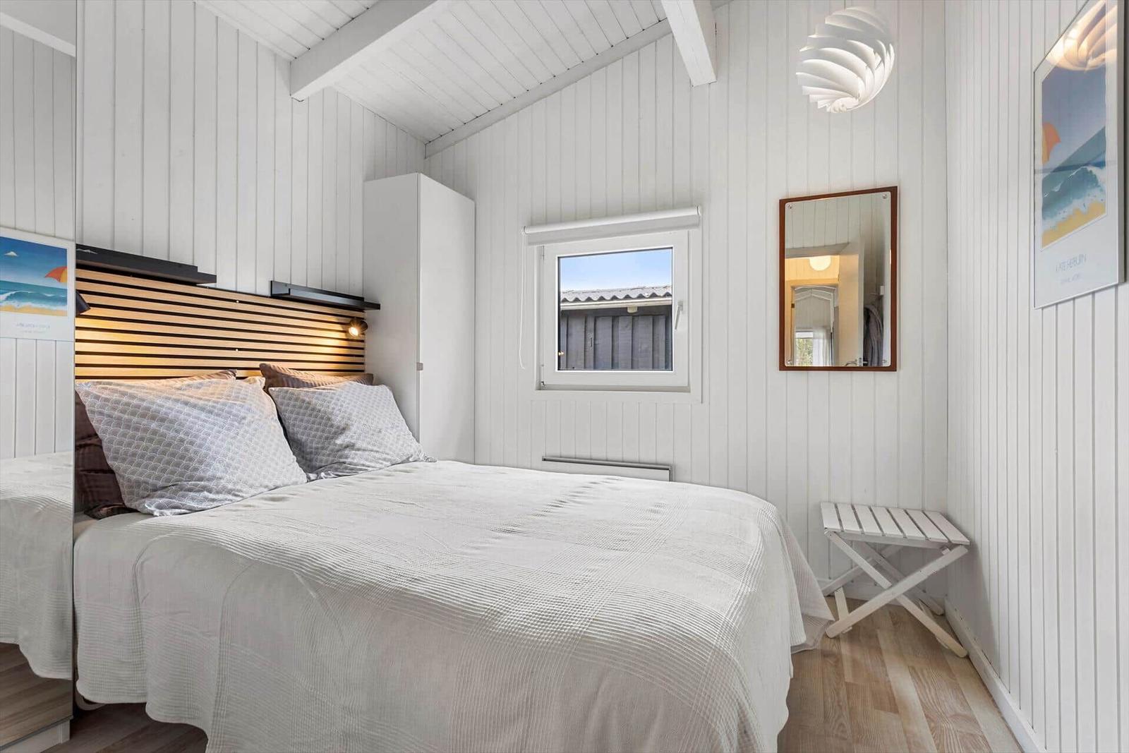 Schlafzimmer mit Bett, Fenster, Spiegel und Stuhl. Wände weiß verkleidet.