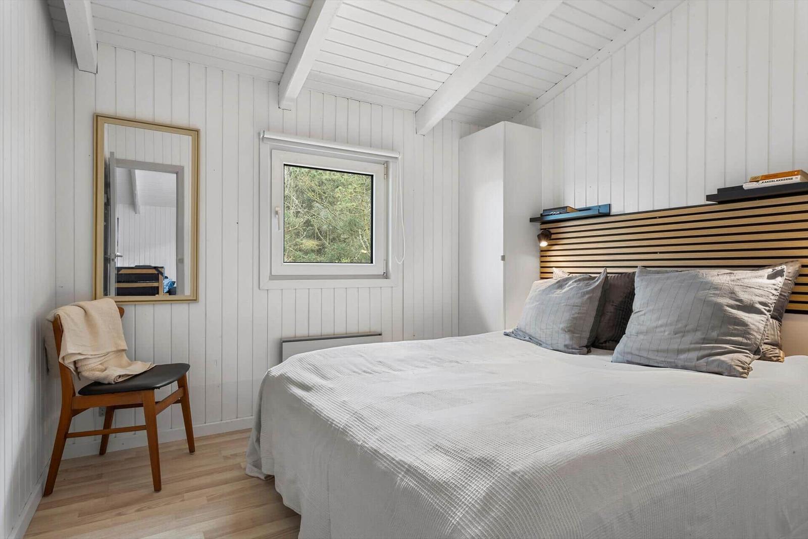 Schlafzimmer mit Bett, Holzstuhl, Spiegel und Fenster zur Natur.