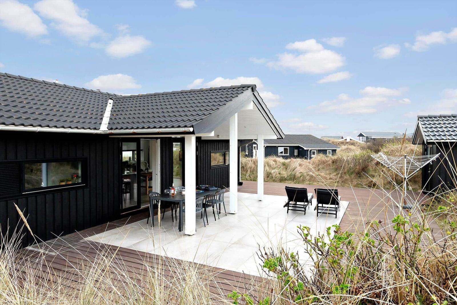 Schwarzes Haus mit Terrasse, Tisch, Stühlen und Liegen. Hintergrund: Dünen und Himmel.
